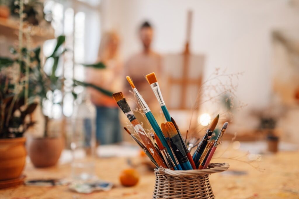 Ein Korb voller Malpinsel in verschiedenen Größen und Farben steht auf einem Tisch in einem künstlerischen Atelier. Im Hintergrund sind unscharf zwei Personen vor einer Staffelei zu erkennen. Die Szene ist hell und von Pflanzen umgeben, was eine kreative und inspirierende Atmosphäre vermittelt.