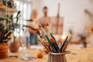 Ein Korb voller Malpinsel in verschiedenen Größen und Farben steht auf einem Tisch in einem künstlerischen Atelier. Im Hintergrund sind unscharf zwei Personen vor einer Staffelei zu erkennen. Die Szene ist hell und von Pflanzen umgeben, was eine kreative und inspirierende Atmosphäre vermittelt.
